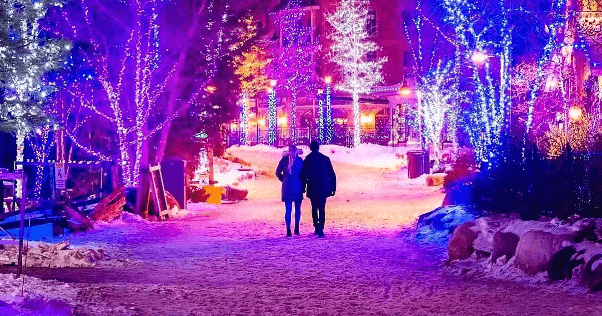 A postcard-esque photo of a couple walking along the Holiday Magic Light Trail. The snow-covered pedestrian streets are glowing in purple and blue lights