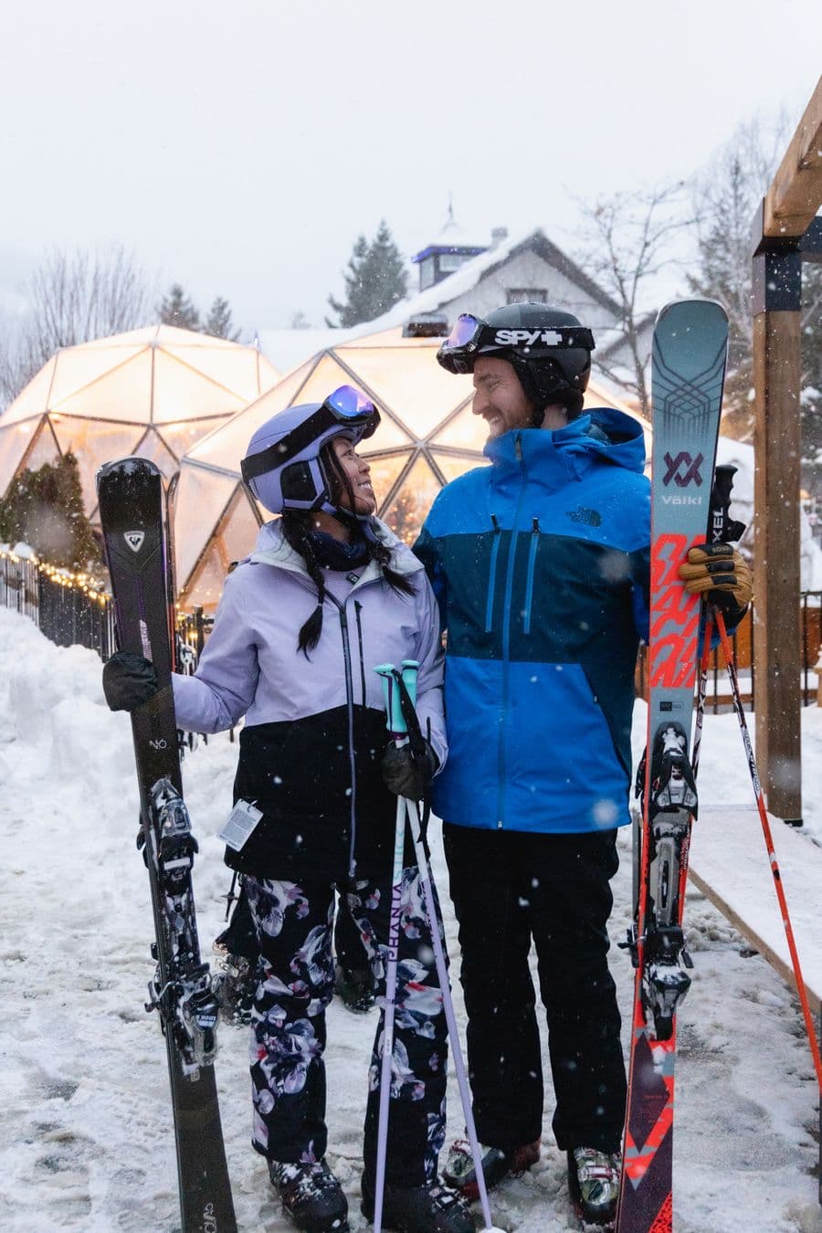 A couple dressed for skiing stare into each other's eyes, smiling, while standing in the Village Events Plaza on a snowy day.