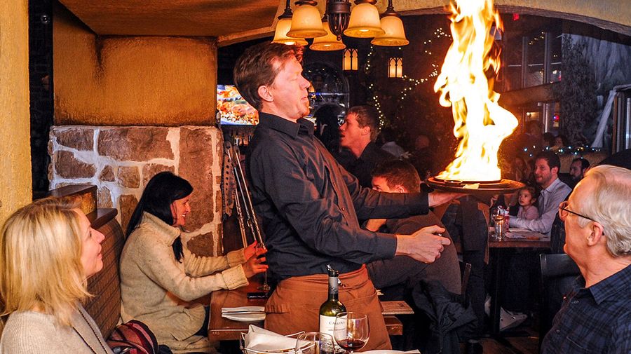 A large flame bursts as a server flambés traditional saganaki tableside at Tholos Greek Restaurant
